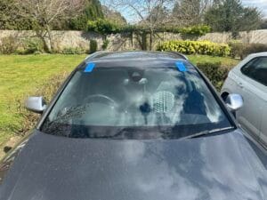 Front view of a grey car with a cracked windshield marked with blue tape, parked in a garden with a fence and greenery in the background
